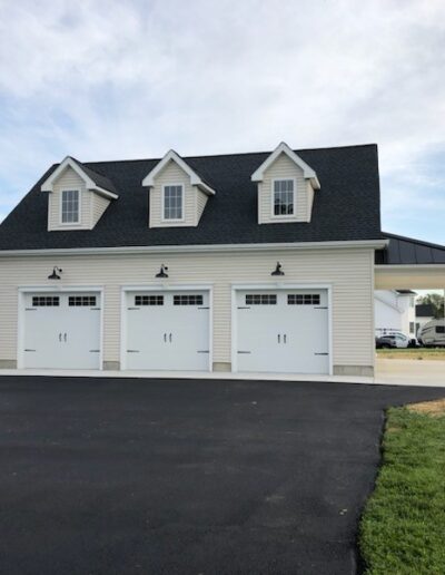 A white three-car garage with a black roof and dormer windows, set on a paved driveway, with grass and trees in the background.